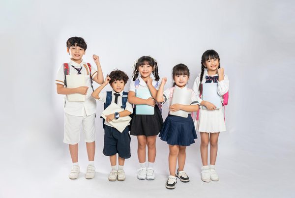 Five young students in school uniforms holding notebooks and backpacks.